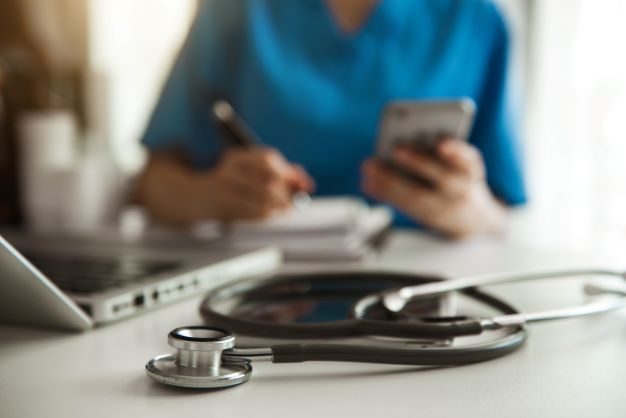 Stethoskop liegt auf dem Tisch, dahinter eine Person in Arztkittel, die ein Handy in der Hand hält