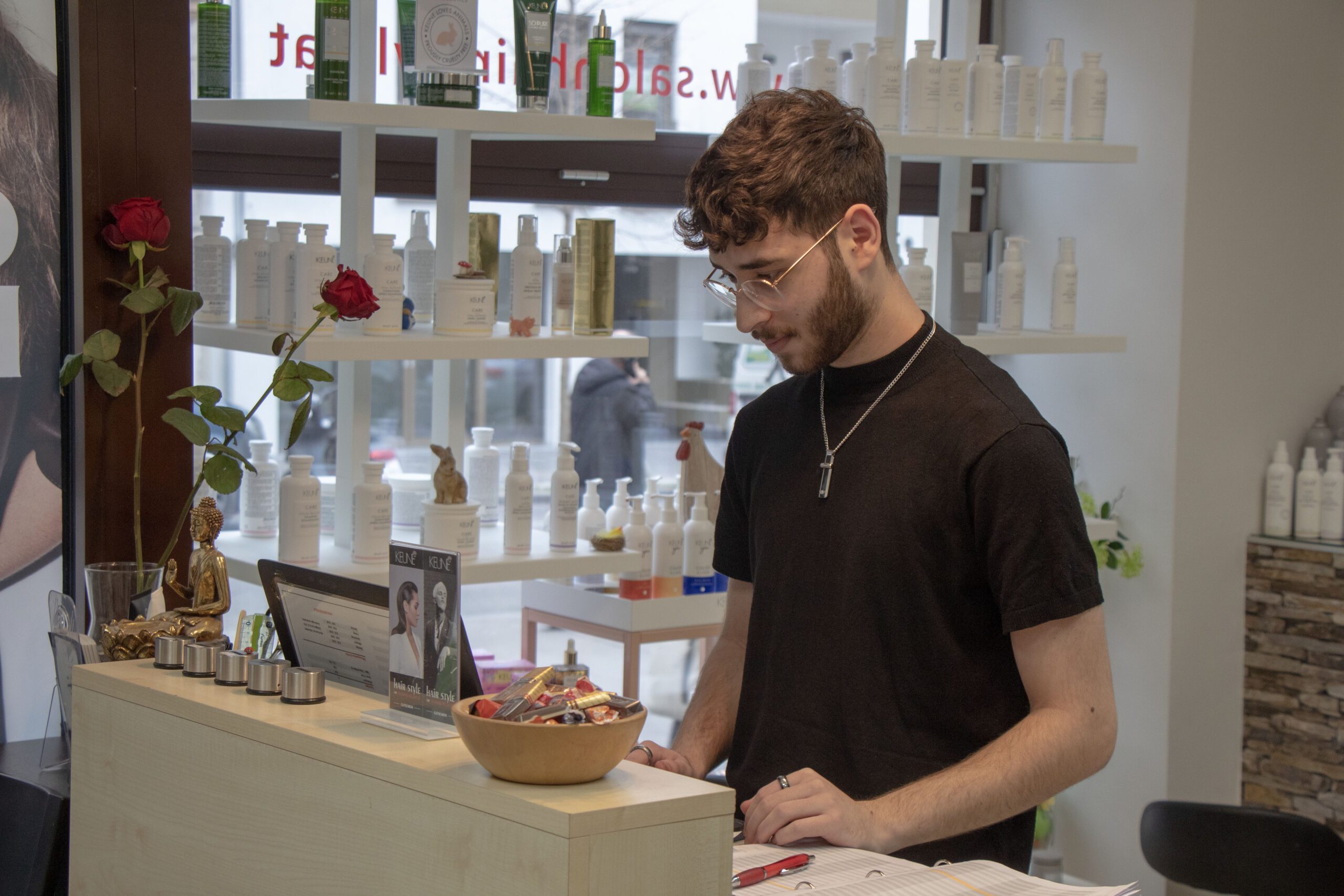Friseurlehrling Aykut Karabas steht am Empfang hinter dem Thresen und blickt in die Kund:innenkartei, neben ihm sind Produkte und 2 Rosen platziert